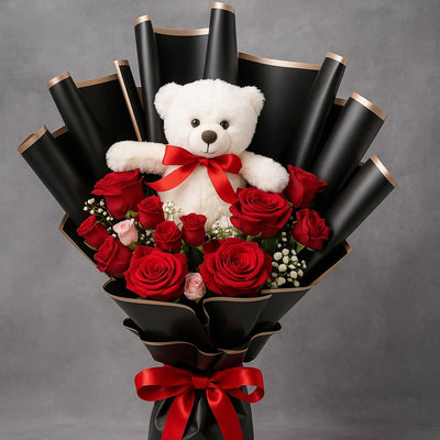 Bouquet of red roses with a white teddy bear wearing a red bow, all wrapped in black paper with gold edges on a gray background.