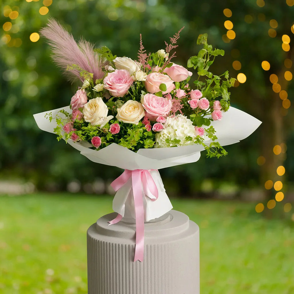 Bouquet of pink and white flowers with greenery on a pedestal against a blurred outdoor background.