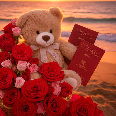 Teddy bear with red and pink roses and red envelopes on a beach at sunset.