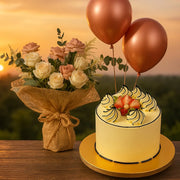 White cake with strawberry decorations, two bronze balloons, and a bouquet of flowers on a wooden table with a sunset background.
