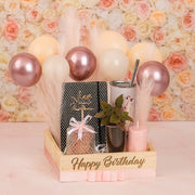 Gift set with balloons, notebook, and other items in a 'Happy Birthday' box against a floral background.