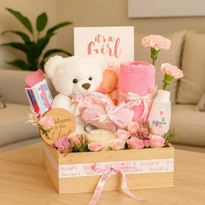 Newborn baby gift basket with teddy bear, baby clothes, and pink flowers on a wooden box.