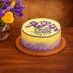 Load image into Gallery viewer, Birthday cake with purple and yellow flowers and &#39;Happy Birthday&#39; text on a wooden table.
