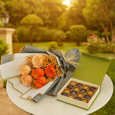Bouquet of orange and peach flowers with a box of chocolates on a table outdoors.