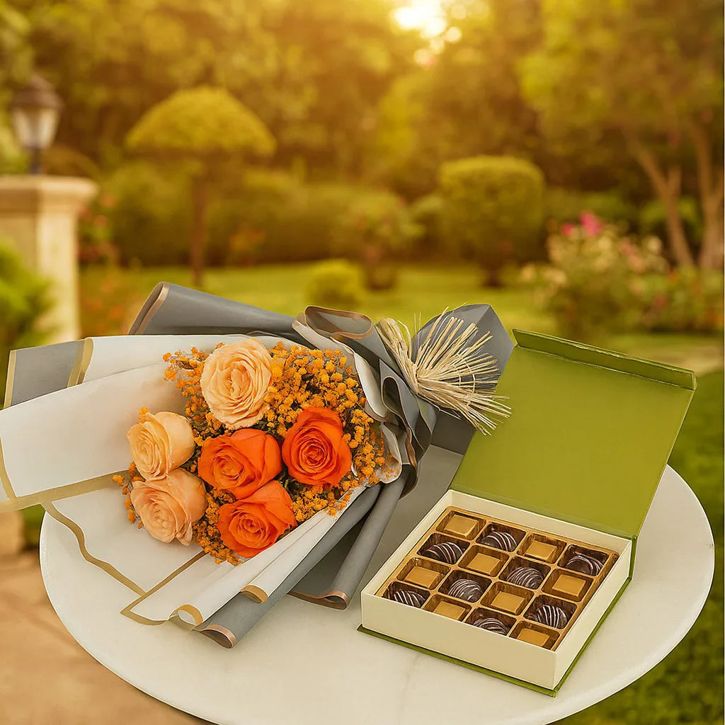 Bouquet of orange and peach flowers with a box of chocolates on a table outdoors.