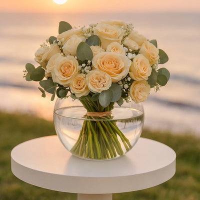 Bouquet of yellow roses in a clear vase on a white table with a sunset background.