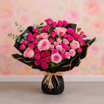 Load image into Gallery viewer, Bouquet of pink and red roses wrapped in black paper with a decorative bow on a floral background.
