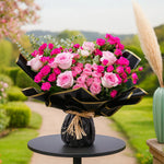 Load image into Gallery viewer, Bouquet of pink and white roses wrapped in black paper on a small round table with a garden background.
