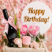 Birthday gift basket with wine, flowers, and 'Happy Birthday!' sign on a pink floral background.
