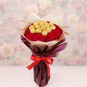 Bouquet of red roses with gold Ferrero Rocher chocolates wrapped in brown paper and a red ribbon on a blurred floral background.