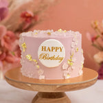 Load image into Gallery viewer, Pink birthday cake with &#39;Happy Birthday&#39; text on a wooden stand against a blurred floral background
