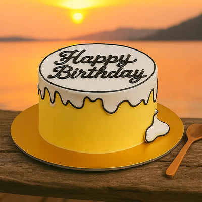 Yellow birthday cake with 'Happy Birthday' on a wooden table against a sunset backdrop