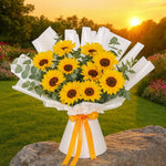 Load image into Gallery viewer, Bouquet of sunflowers wrapped in white paper with a yellow ribbon, set against a scenic background with trees and sunset.
