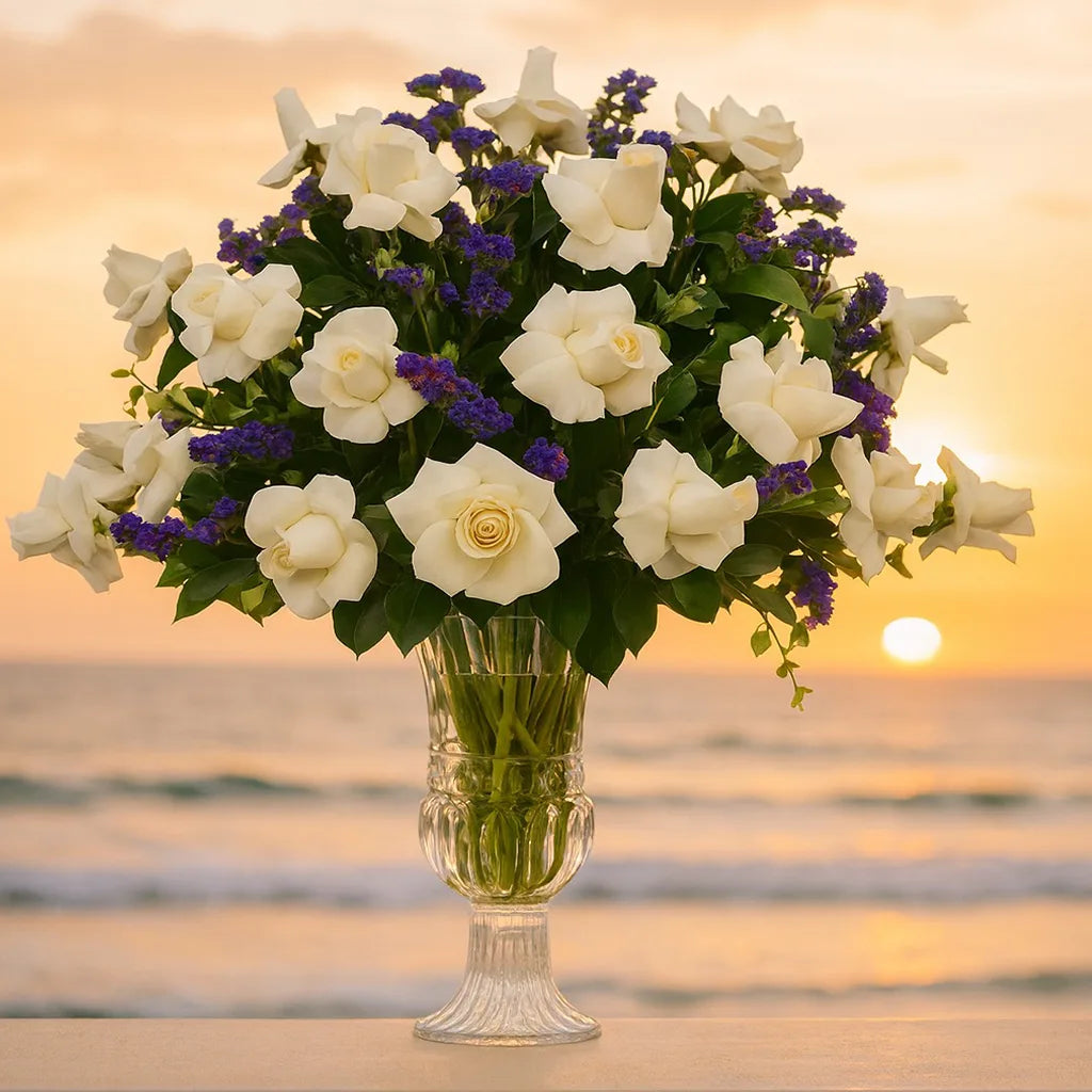 Bouquet of white and purple flowers in a clear vase against a sunset beach backdrop